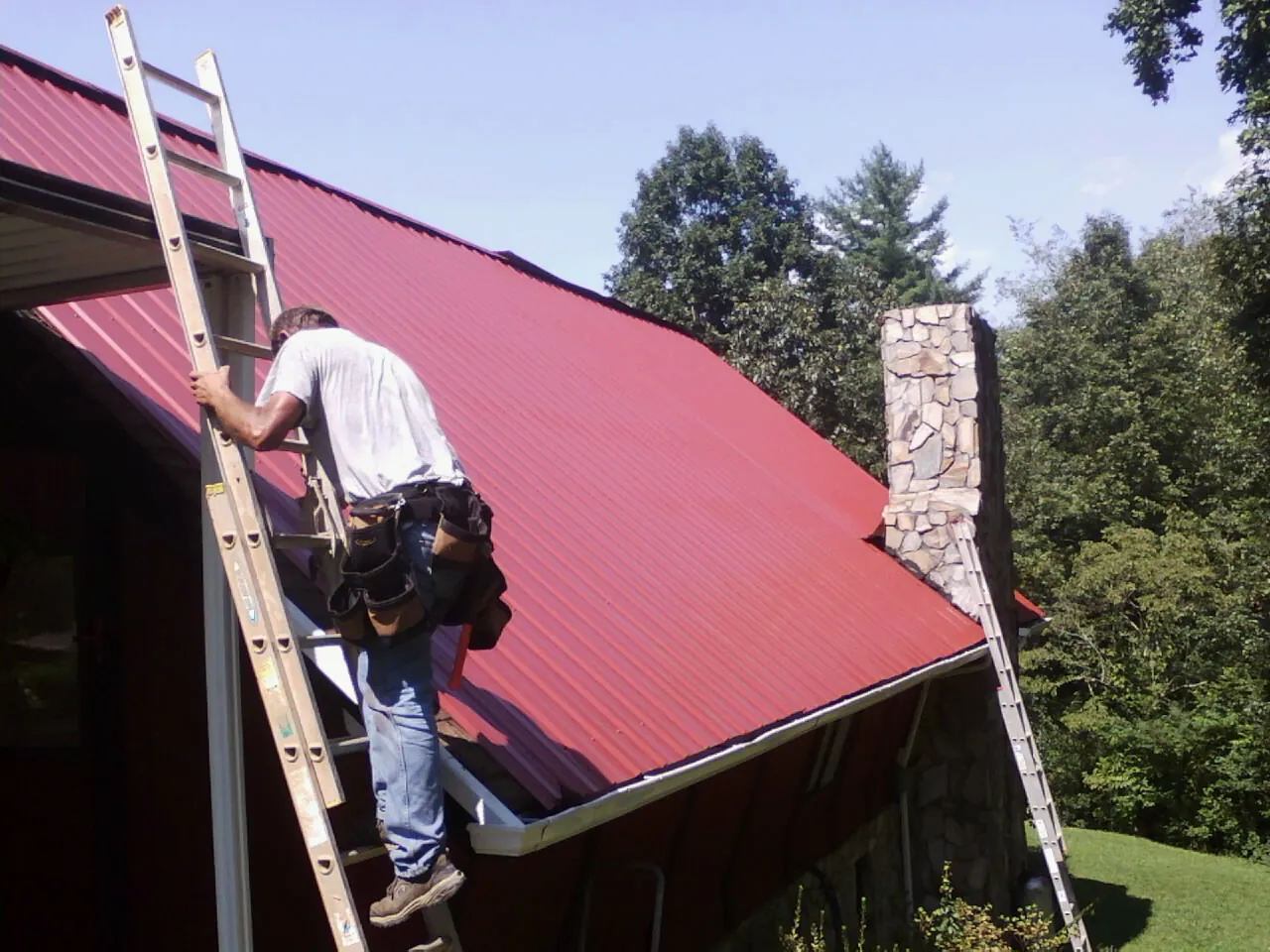 Roofer on a ladder beside a red metal roof and stone chimney — Soffit & Fascia Repair by Roofing Brothers in Custer